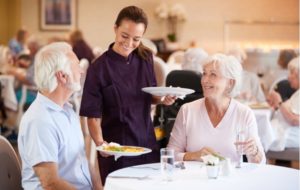 A senior living chef serving two seniors at a dinner table in the dining room of their independent living community