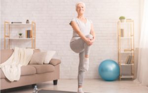 A senior woman in her suite performing a flamingo stand, raising her knee up to her chest to strengthen her balance