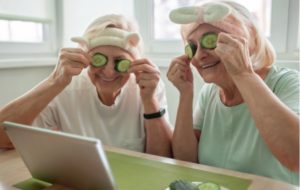 Two senior women having fun, holding cucumber slices over their eyes while they follow instructions on a tablet to do a face mask