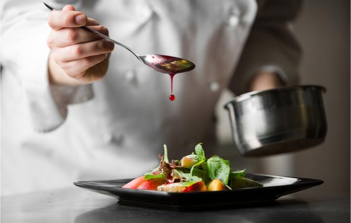 A chef adding the final touches to a meal, drizzling a sauce on the top of some green leaves and potatoes