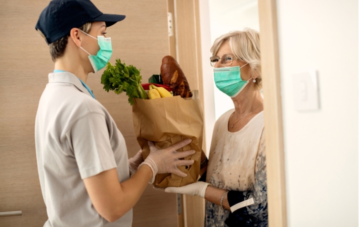 A female courier delivering a brown paper bag of groceries to a senior woman in her doorway