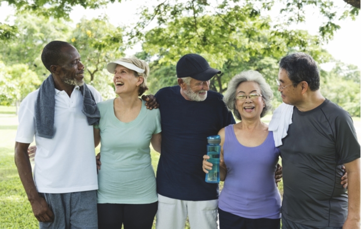 A group of five seniors in active clothing, standing with their arms around each other in a park, smiling after they just exercised