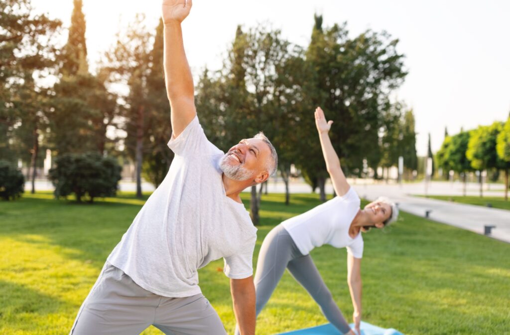Two older adults raise their arms to the sky while stretching outdoors in a beautiful sunlit park