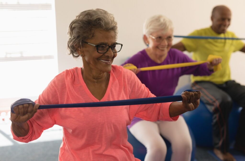 Three older adults in a row grin while stretching resistance bands and sitting on medicine balls together during a fitness class in senior living