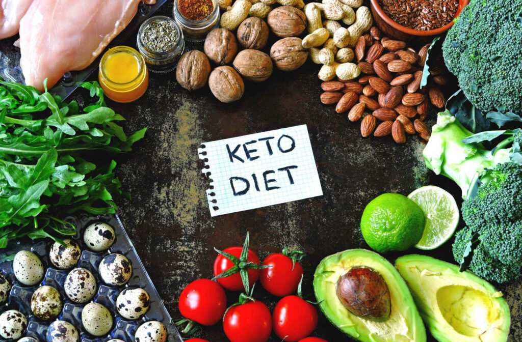 A variety of food items placed on a table that make up a keto diet like avocados, tomatoes, almonds, eggs, chicken and nuts.