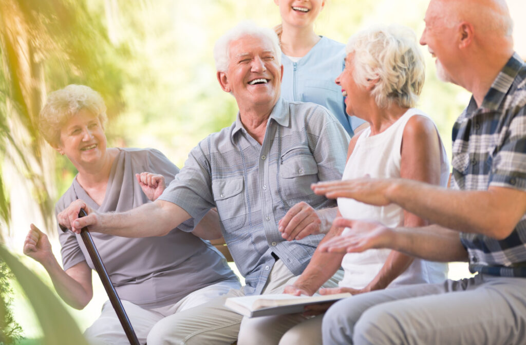 A senior man with a cane smiles and laughs while sitting outside with a nurse and other seniors