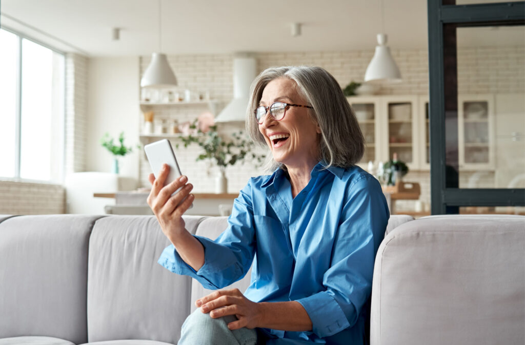 A senior woman sitting on a couch, smiling and holding a smartphone in her right hand for a video call.