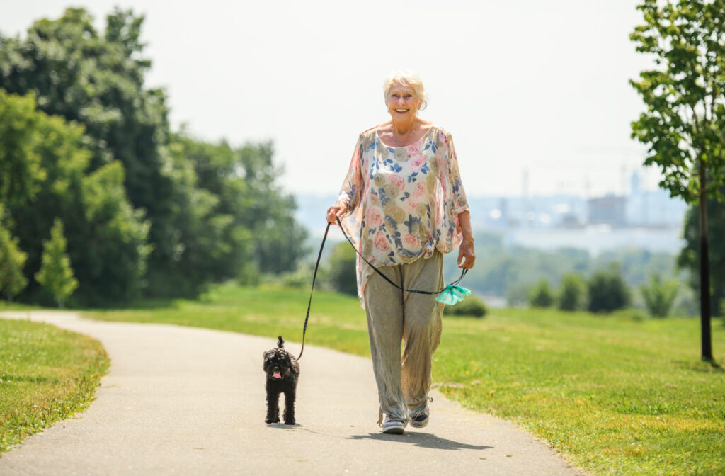 A senior woman smiling and walking her pet poodle in a park.