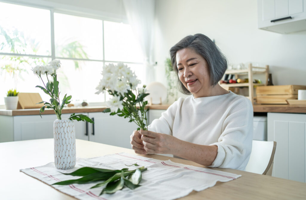 An older adult woman smiling and putting flowers in a vase.