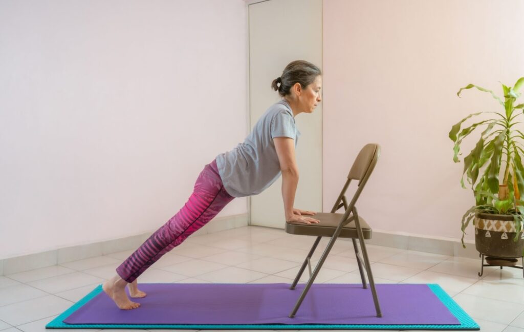 A senior uses a folding chair to support themselves in a yoga pose during a chair yoga class at their senior living community.