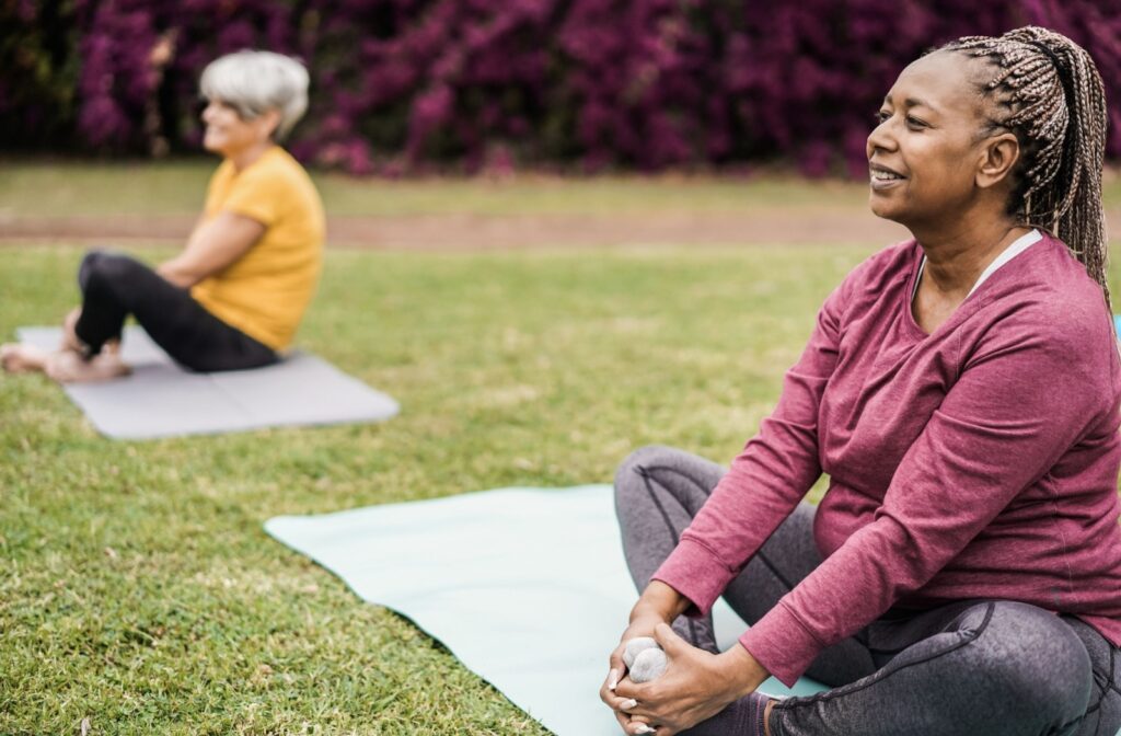 Two older adults sit in a grassy area outside on yoga mats. They gently pull their feet towards their hips in a deep stretch.