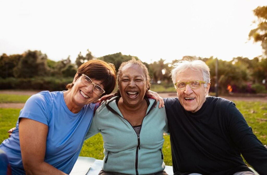 Three older adults sitting outside on a sunny day while smiling and laughing with arms around each other