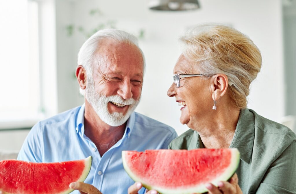 Two older adults laughing indoors while holding large slices of watermelon together