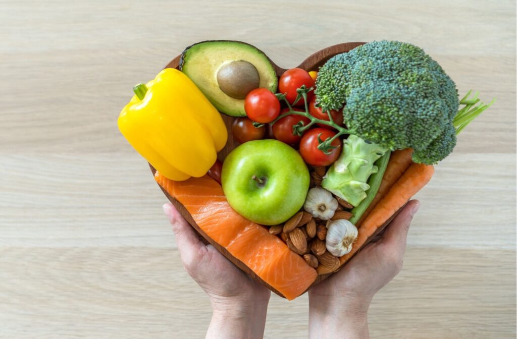 Over a wooden table, a pair of hands holds up a heart-shaped bowl full of heart-healthy foods like salmon, avocados, and nuts