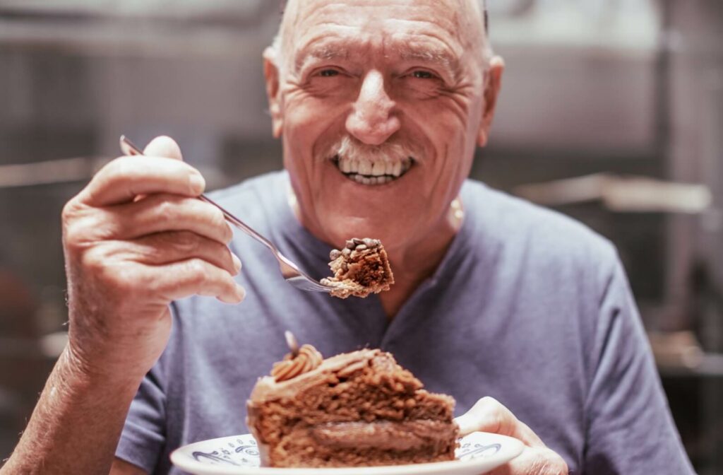 A smiling older adult holds a bite of dark chocolate cake on a fork, while holding a plate with the rest of the cake slice