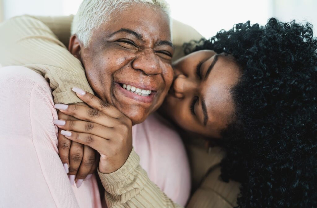 An adult child hugs and kisses their smiling senior parent on the cheek