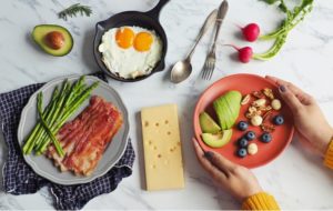 A spread of various keto-friendly foods including bacon, eggs, avocado, nuts, berries, cheese, and asparagus
